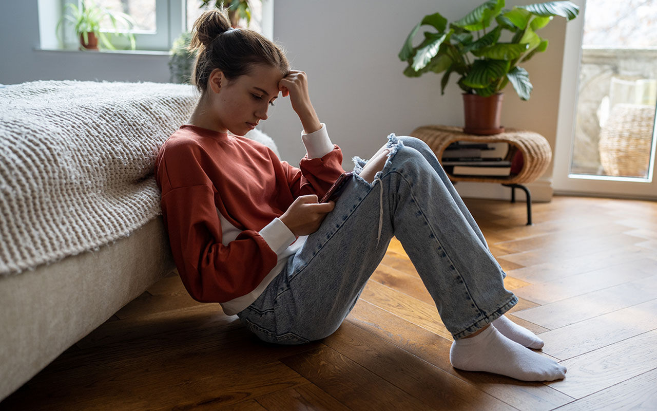 En person sitter på golvet vid en säng och använder en mobiltelefon.