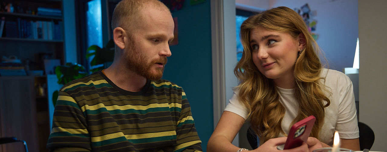 Två personer sitter vid ett matbord och tittar på en mobiltelefon.