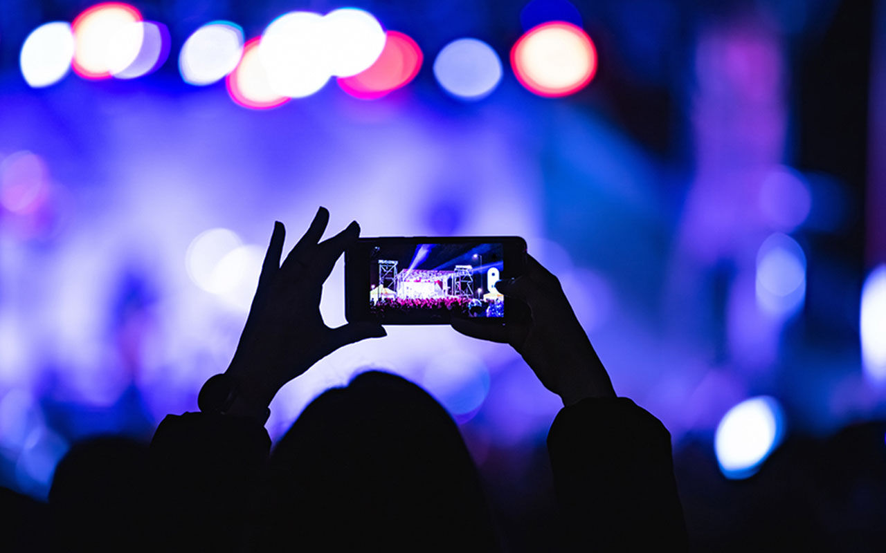 En person håller upp en smartphone och fotograferar en scen med starka färgade ljus på en konsert eller ett evenemang, med suddig publik och scen i bakgrunden.